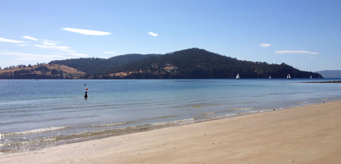People swimming in calm water at Nebraska Beach, Bruny Island, with sailing boats in the background.
